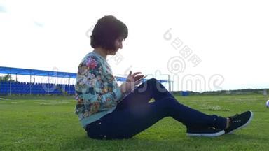 在阳光明媚的日子里，体育场草坪上一位迷人的年轻女子使用智能手机。